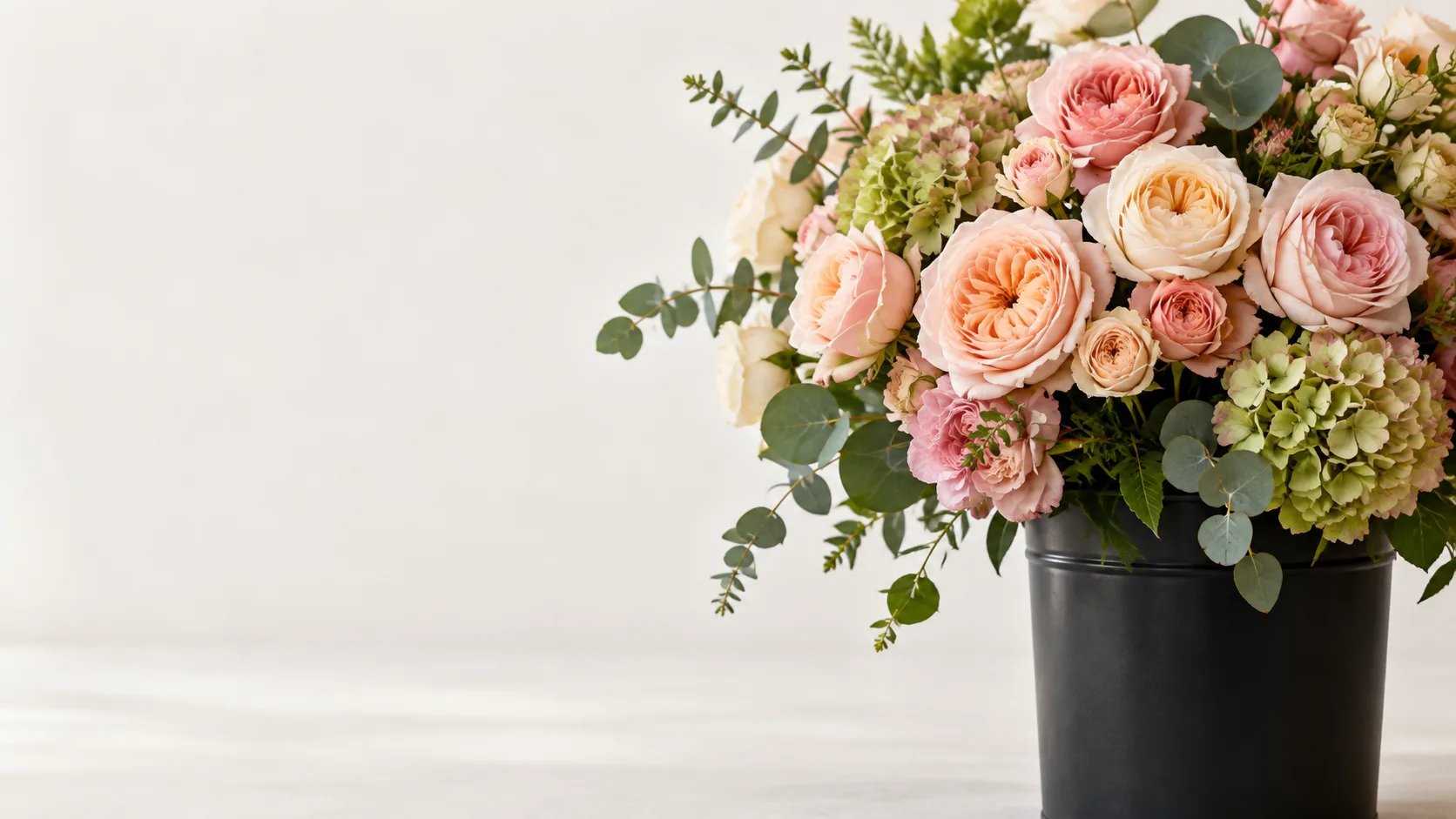 Rosas de jardín en tonos melocotón y rosa, hortensia verde y eucalipto en un cubo negro